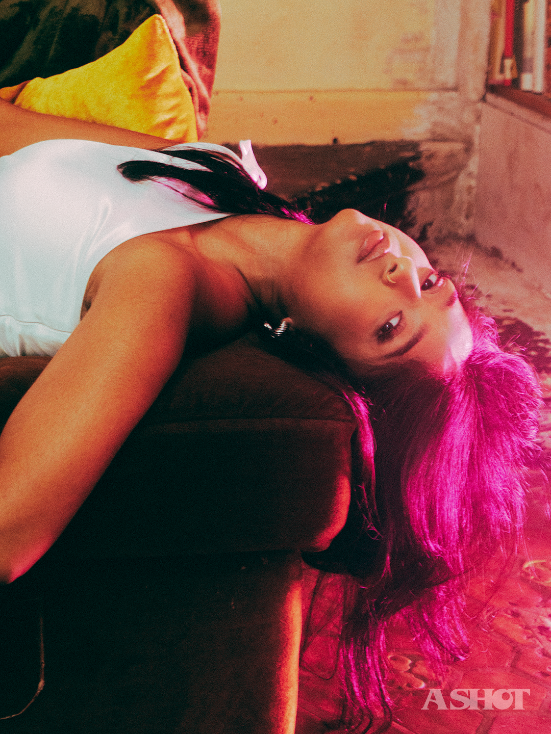 Close up of Maitreyi Ramakrishnan laying across a couch with her hair handing off the edge while looking at the camera